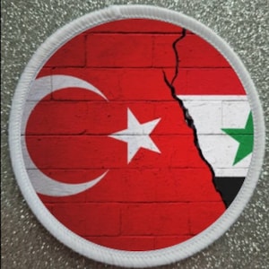 May include: A white circular patch with a red brick wall background. The wall is cracked down the middle, with the Turkish flag on the left and the Syrian flag on the right.