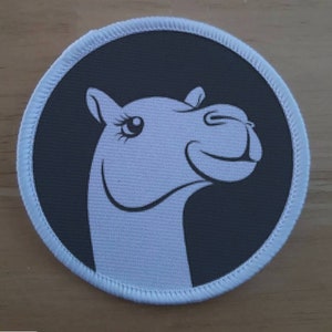 May include: A white camel head with a black outline and a winking eye is printed on a round, gray fabric patch with white stitching.