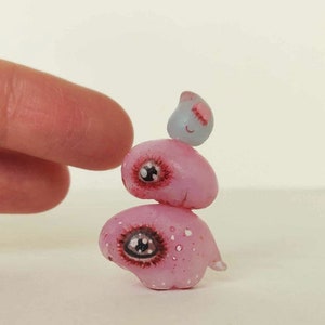 May include: A stack of three pink and gray clay figures with painted eyes and details. The figures are stacked on top of each other, with the smallest figure on top. The figures are handmade and have a whimsical, cartoonish style.