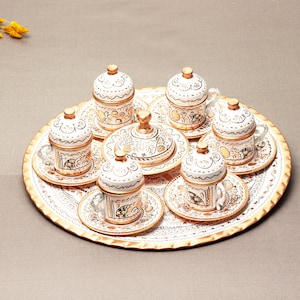 Puede incluir: Un juego de café turco blanco y dorado con seis tazas y platillos, un azucarero y una bandeja. Las tazas y los platillos tienen intrincados diseños florales.