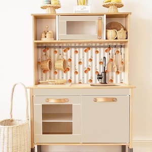 Könnte beinhalten: Eine Spielküche aus Holz in hellem Beige, mit Mikrowelle, Spüle und Stauraum. Der Küchenhintergrund hat ein Streifenmuster mit Orangenfrüchten. Enthält Hängekörbe und Holzutensilien.