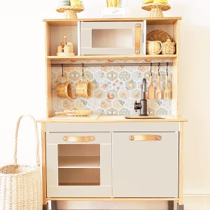May include: A wooden play kitchen with a white countertop, a gray oven, a black sink, and a white cabinet with a glass door. The kitchen has a backsplash with a honeycomb pattern and floral accents. There are hooks on the backsplash for hanging utensils.