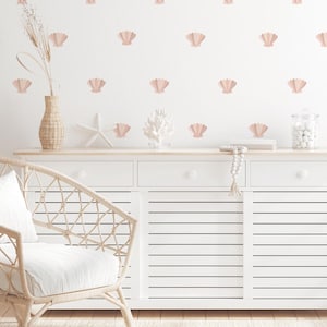 Op de afbeelding: Een witte commode met drie lades en een witte muur met roze schelp stickers. De commode heeft een houten blad en een witte rieten stoel met een wit kussen.