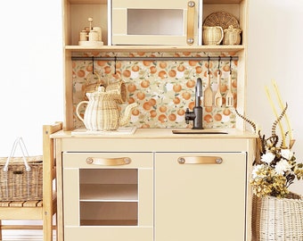 Può includere: Una cucina giocattolo in legno con finitura marrone chiaro. La cucina ha un forno a microonde, un lavandino e due armadietti con ante in vetro. Il paraschizzi è decorato con un motivo floreale arancione e bianco. "Cucina, maniglie e decorazioni non incluse" è stampato sotto la cucina.