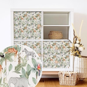 Könnte beinhalten: Weißer Schrank mit Schubladen- und Türfronten im Dschungel-Design. Das Design zeigt Giraffen, Zebras, Elefanten und Papageien inmitten üppigen Grüns. Ein rundes Detail befindet sich auf den Schubladen.