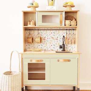 Pode incluir: Uma cozinha de brinquedo de madeira verde claro com micro-ondas, pia e armários. Possui um backsplash com estampa floral, cestas de vime e utensílios de madeira. Uma cesta de vime está à esquerda.