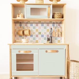 May include: A light blue wooden play kitchen with a white tiled backsplash, a brown leather handle, and a black faucet. The kitchen has a microwave, a sink, and two cabinets with shelves.