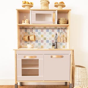 May include: A wooden toy kitchen with a light pink finish, featuring a microwave, sink, and cabinets. It includes a checkered backsplash, hanging utensils, and small woven baskets. The handles are tan leather.