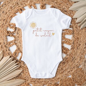 May include: White baby bodysuit with short sleeves. The bodysuit has a sun graphic and the text "petit rayon de soleil" in a script font. A small gold heart is also included in the design. The bodysuit is on a woven mat.