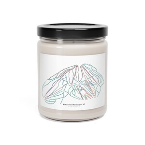 Op de afbeelding: Een witte kaars in een glazen pot met een zwart deksel. Het etiket op de pot heeft een kleurrijke lijntekening van een berg met de tekst "Stratton Mountain, VT 43.1304° N, 73.0601° W".