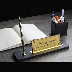 May include: A black leather desk nameplate with a gold nameplate insert. The nameplate reads "Adam Smith General Manager". A gold pen is in a holder on the desk.