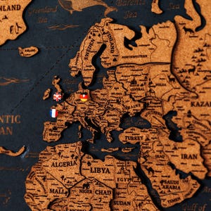 Peut inclure: Une carte en bois détaillée de l'Europe et des environs, avec les contours et les noms des pays. La carte est faite d'un matériau brun clair, monté sur un fond bleu foncé. De petits drapeaux épinglés marquent divers endroits.