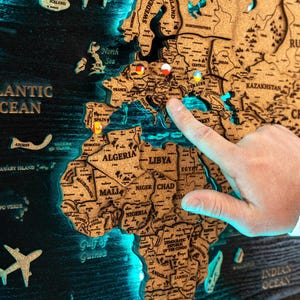 May include: A wooden world map with countries and oceans carved into it, illuminated by blue LED lights. A hand points to a location on the map, with pins marking various places. The words "Atlantic Ocean" are visible.
