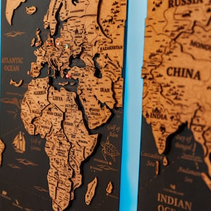 Peut inclure: Une carte du monde en bois détaillée, avec les contours et les noms des pays gravés sur un fond sombre. La carte comprend l'océan Atlantique, la Chine et l'Inde. Des épingles sont présentes. C'est une pièce décorative.