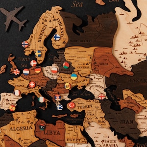 Peut inclure: Carte du monde en bois de l'Europe, avec des contours de pays en différentes nuances de marron et de beige. De petits drapeaux épinglés marquent différents pays. Un avion en bois et les mots "Océan Atlantique" sont également visibles.