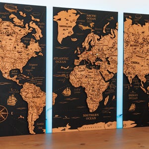 Peut inclure: Une carte du monde en bois en trois panneaux avec un fond noir. La carte est détaillée avec les contours des pays et les noms des océans en marron clair. Une rose des vents est présente sur le panneau de gauche. Les panneaux sont affichés sur une surface en bois.