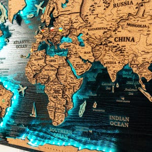 Peut inclure: Une carte du monde en bois détaillée, avec des contours de pays découpés au laser et des océans. Les océans sont mis en valeur par un éclairage LED bleu. Des avions et des voiliers miniatures décorent la carte, ainsi que des épingles.