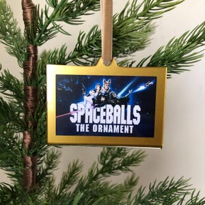 May include: A gold-colored Christmas ornament featuring a scene from the movie Spaceballs. The ornament has a white background with a blue and black image of the characters from the movie. The text "Spaceballs The Ornament" is printed in white on the ornament.