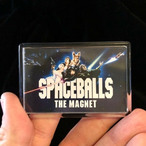 May include: A clear plastic frame with a black background featuring a movie poster for the film "Spaceballs". The poster shows the main characters from the film, including Lone Starr, Barf, and Princess Vespa. The text "Spaceballs" is in white letters with a red laser beam shooting across the image. The text "The Magnet" is below the title in white letters.