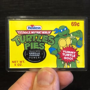 Puede incluir: Una caja amarilla de tartas de tortuga Hostess Teenage Mutant Ninja Turtles con una imagen de dibujos animados de una tortuga verde con un pañuelo azul y sosteniendo una tarta. La caja dice "Teenage Mutant Ninja Turtles Pies" y "Filled with Vanilla Puddin' Power!" y "Yummy Turtle Goo!" La caja cuesta 69 céntimos y pesa 5 onzas.