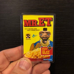May include: A yellow cereal box with the words "MR.ET" in large, black letters. The box features an image of an alien wearing a gold chain and a black hat, holding a bowl of cereal. The text "PITY THE FOOL WHO DON'T PHONE HOME!!" is printed below the alien's image. The box also includes the Quaker logo and a General Mills logo. The text "FREE TOY MR.ET INSIDE" is printed in a red box on the right side of the box.