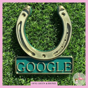 May include: A gold horseshoe with a green and gold sign that says "GOOGLE" on it. The sign is on a green grassy background. The horseshoe is a symbol of good luck.