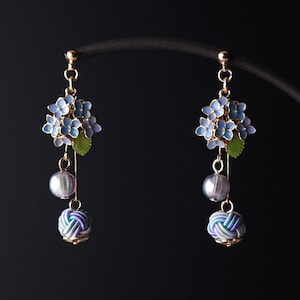 May include: A pair of gold-toned earrings featuring blue floral clusters, green leaves, and dangling elements. Each earring has a pearl and a decorative knot in shades of blue and white. The earrings are displayed against a dark background.