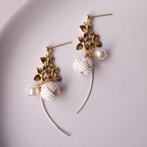 May include: A pair of gold-coloured earrings with a floral design and white knotted details. Each earring has a small pearl and a curved gold wire. The earrings are presented on a white, oval-shaped plate.