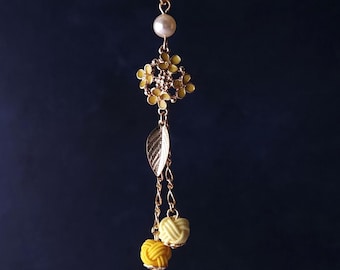 May include: A gold-coloured hair stick featuring a floral and leaf design. A pearl sits at the top, with a cluster of small flowers, a leaf, and two yellow and green decorative balls hanging below. The background is a dark blue.