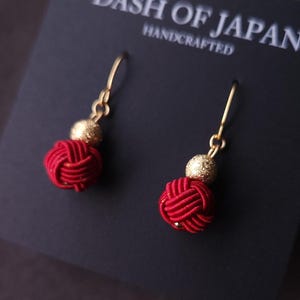 May include: A pair of handcrafted red and gold earrings. Each earring features a red knot design and a gold bead, suspended from gold-colored hooks. The earrings are displayed on a black card with the text "DASH OF JAPAN HANDCRAFTED".