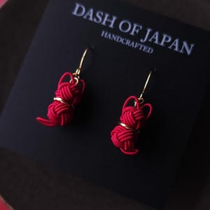 May include: Red handcrafted earrings with gold-colored accents, presented on a black card with the text "DASH OF JAPAN HANDCRAFTED". The earrings are displayed on a dark brown plate against a red background.