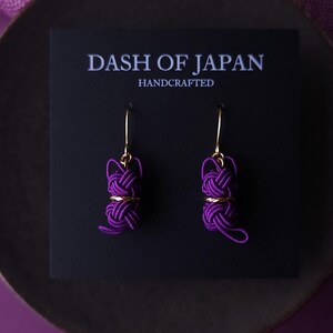 May include: A pair of purple cord knot earrings with gold-toned hooks attached to a black display card. The card features the white text DASH OF JAPAN HANDCRAFTED. The earrings consist of intricate, tightly wound purple cord knots with gold accents.