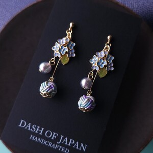 May include: A pair of handcrafted earrings featuring blue and gold floral clusters, gray pearl drops, and patterned ball accents. The earrings are displayed on a black card with the text "DASH OF JAPAN HANDCRAFTED".