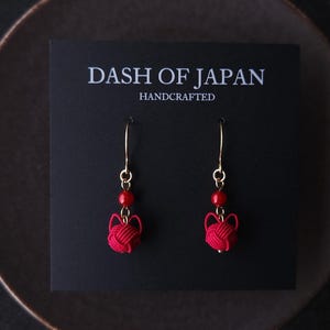 May include: Red knot earrings with gold-colored hooks and red beads. The earrings are displayed on a black card with the text "DASH OF JAPAN HANDCRAFTED". The earrings are presented on a dark brown plate.
