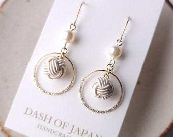 May include: A pair of gold-tone drop earrings featuring a white cord knot suspended within a textured gold circle, accented by a single white pearl bead. The earrings are displayed on a white card printed with the text DASH OF JAPAN HANDCRAFTED.