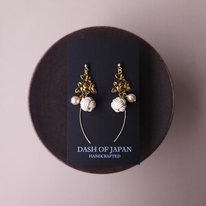 May include: Gold floral earrings with white knotted details and a pearl drop. The earrings are presented on a black card with the text "DASH OF JAPAN HANDCRAFTED". The earrings are on a dark brown plate.
