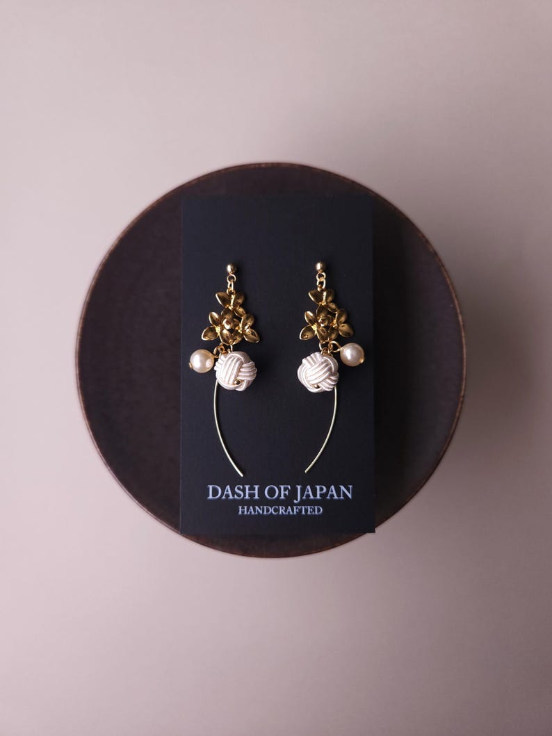 May include: Gold floral earrings with white knotted details and a pearl drop. The earrings are presented on a black card with the text "DASH OF JAPAN HANDCRAFTED". The earrings are on a dark brown plate.