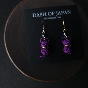 May include: A pair of purple cord earrings featuring a knotted design with gold-tone accents and hooks. The earrings are displayed on a black card printed with the text DASH OF JAPAN HANDCRAFTED in white lettering.