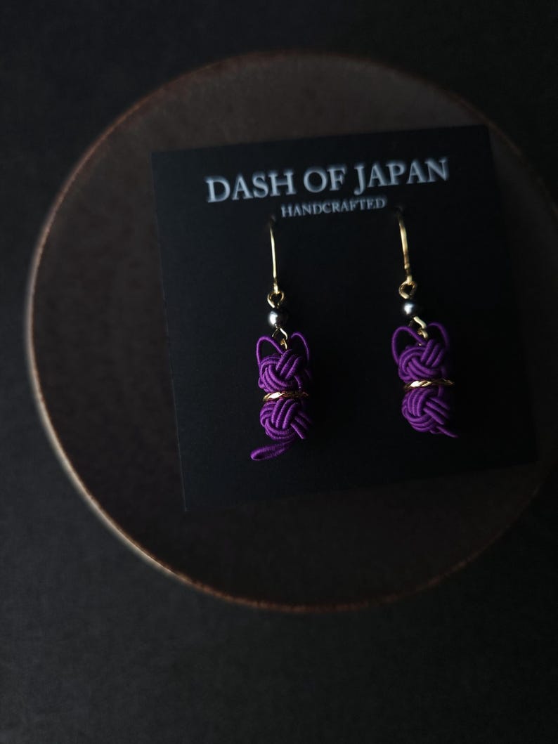 May include: A pair of purple cord earrings featuring a knotted design with gold-tone accents and hooks. The earrings are displayed on a black card printed with the text DASH OF JAPAN HANDCRAFTED in white lettering.