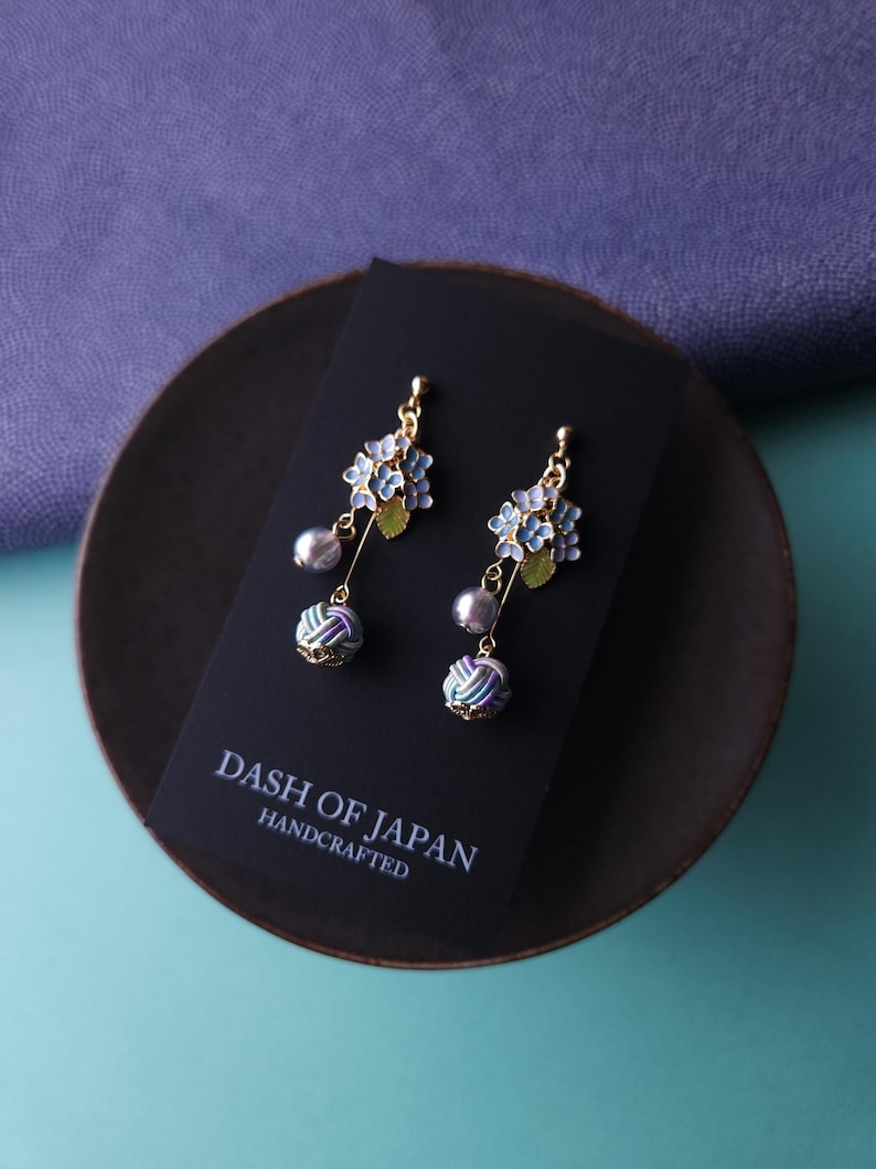 May include: A pair of handcrafted earrings featuring blue and gold floral clusters, gray pearl drops, and patterned ball accents. The earrings are displayed on a black card with the text "DASH OF JAPAN HANDCRAFTED".