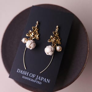 May include: Handmade earrings featuring gold-coloured floral elements, white knotted designs, and pearl drops. The earrings are presented on a black card with the text "DASH OF JAPAN HANDCRAFTED".