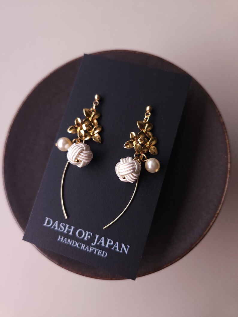 May include: Handmade earrings featuring gold-coloured floral elements, white knotted designs, and pearl drops. The earrings are presented on a black card with the text "DASH OF JAPAN HANDCRAFTED".