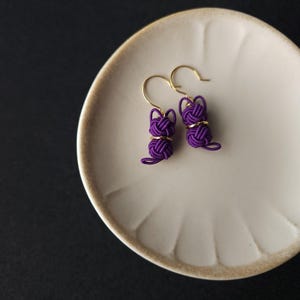 May include: A pair of purple earrings featuring a cat-shaped design crafted from knotted cord. Each earring includes a gold-toned metal hook and a small gold-colored ring accent positioned between the knotted head and body of the cat figure.