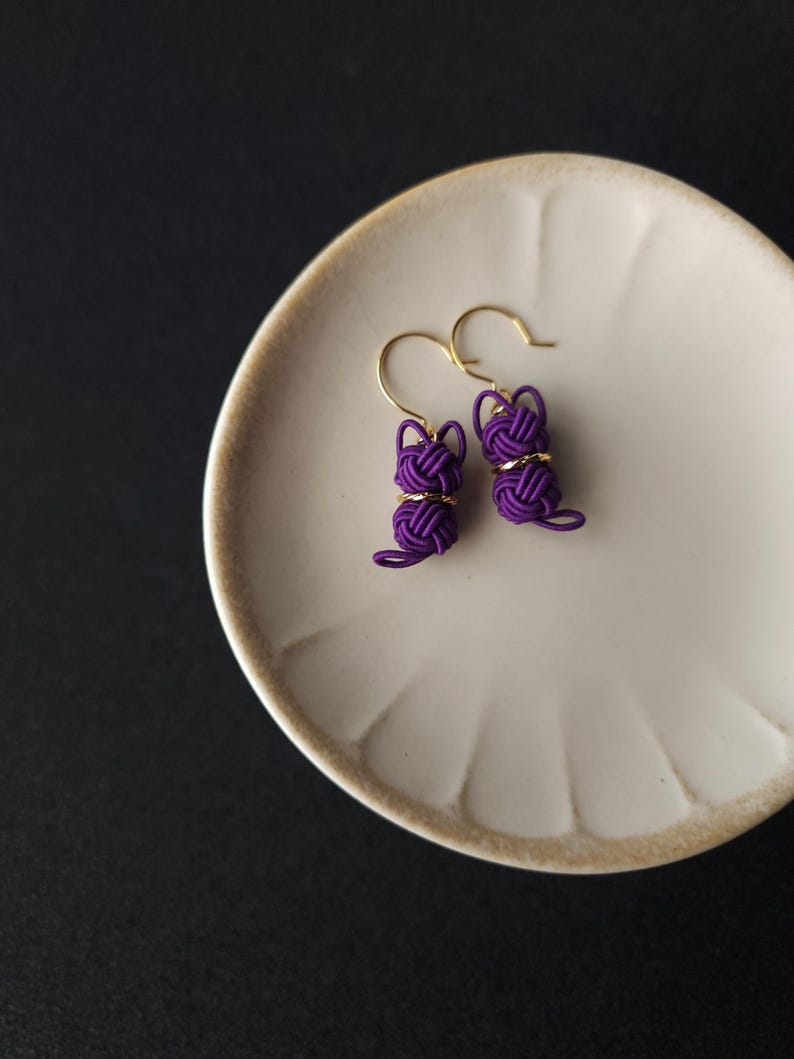 May include: A pair of purple earrings featuring a cat-shaped design crafted from knotted cord. Each earring includes a gold-toned metal hook and a small gold-colored ring accent positioned between the knotted head and body of the cat figure.