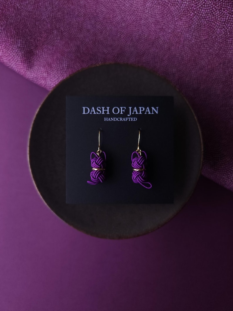 May include: A pair of purple cord knot earrings with gold-toned hooks attached to a black display card. The card features the white text DASH OF JAPAN HANDCRAFTED. The earrings consist of intricate, tightly wound purple cord knots with gold accents.