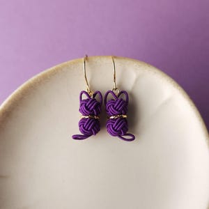 May include: A pair of purple cord earrings shaped like cats, featuring intricate knotted detailing and small gold-toned metal rings. The earrings hang from thin gold-toned hooks, showcasing a distinct feline silhouette created from woven cord.