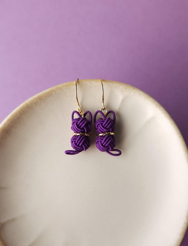 May include: A pair of purple cord earrings shaped like cats, featuring intricate knotted detailing and small gold-toned metal rings. The earrings hang from thin gold-toned hooks, showcasing a distinct feline silhouette created from woven cord.