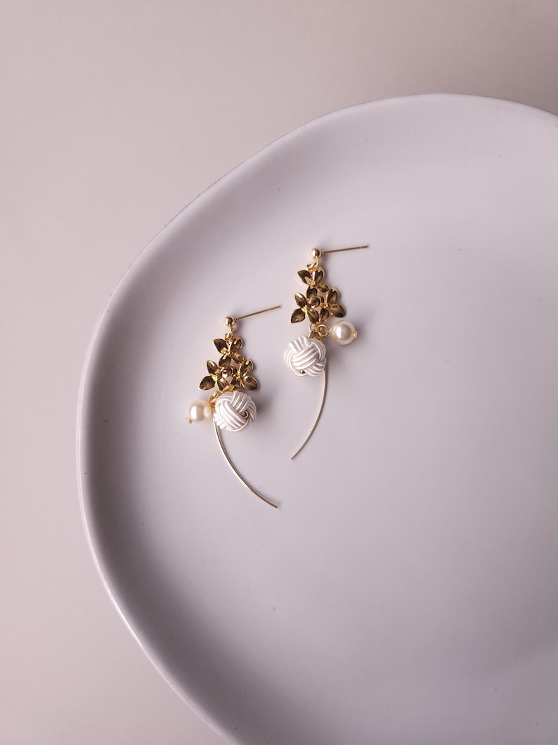 May include: A pair of gold-coloured earrings with a floral design and white knotted details. Each earring has a small pearl and a curved gold wire. The earrings are presented on a white, oval-shaped plate.
