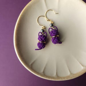 May include: A pair of dangle earrings featuring purple cord knotted into a rabbit shape, accented with a small metallic bead and a gold-toned ring. The earrings hang from gold-toned hooks, showcasing intricate cord work and a playful design.