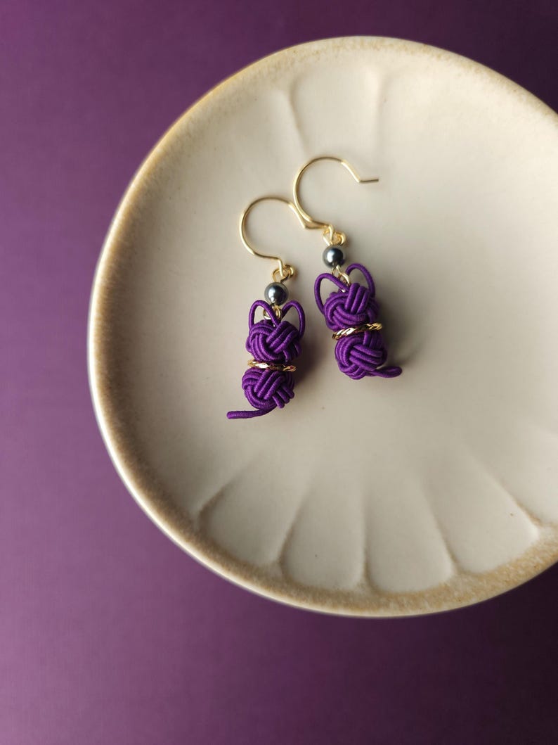 May include: A pair of dangle earrings featuring purple cord knotted into a rabbit shape, accented with a small metallic bead and a gold-toned ring. The earrings hang from gold-toned hooks, showcasing intricate cord work and a playful design.